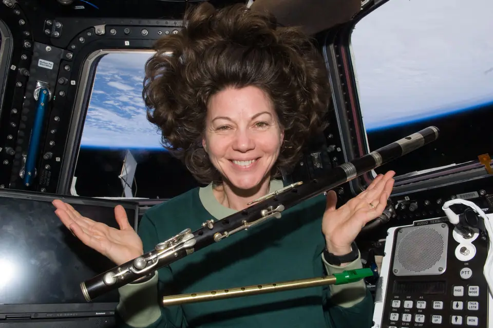 Una astronauta en la cúpula de la Estación Espacial Internacional, con el pelo flotando y una flauta suspendida en el aire; por las ventanas se ve la Tierra