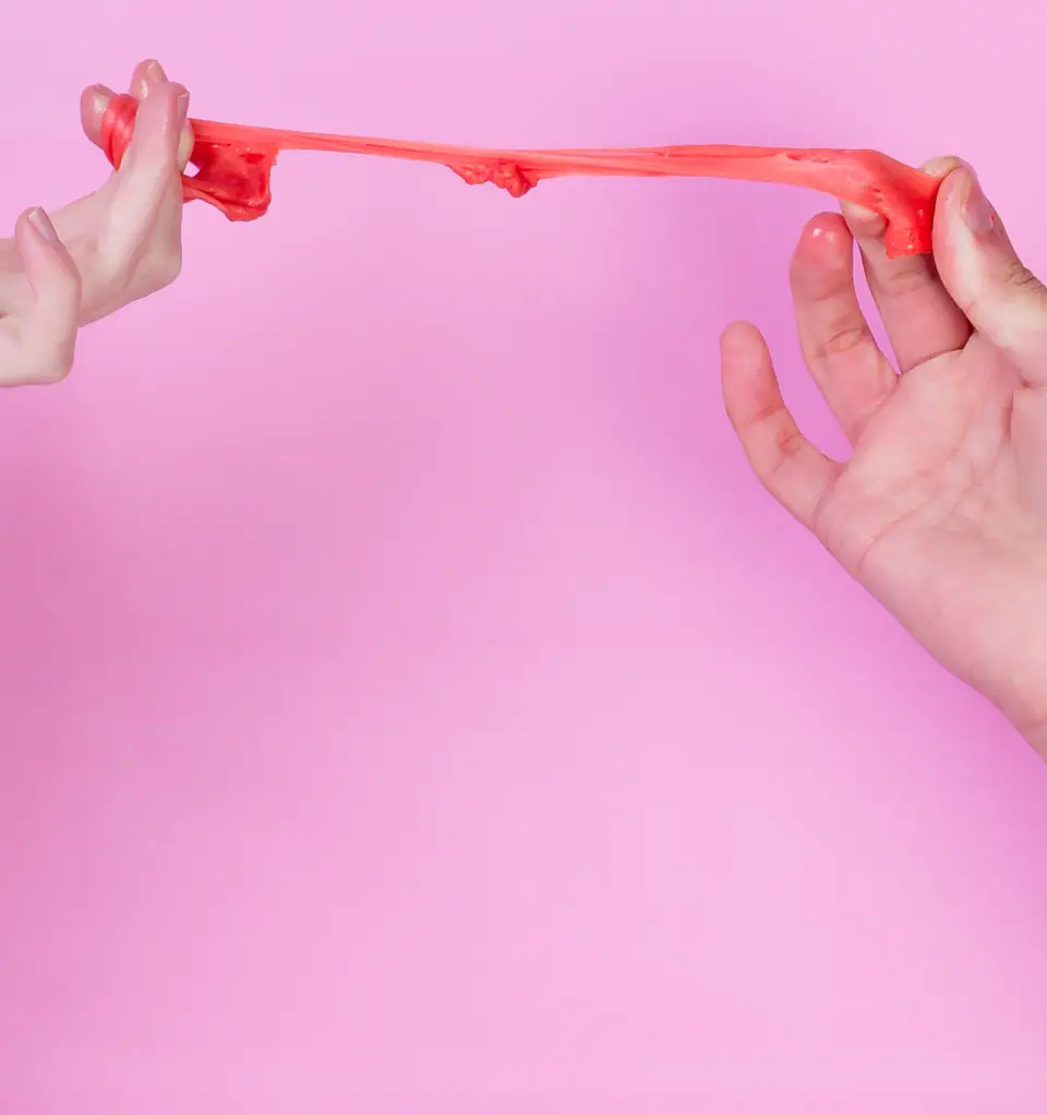 Dos manos estiran un chicle rosado masticado sobre un fondo rosa