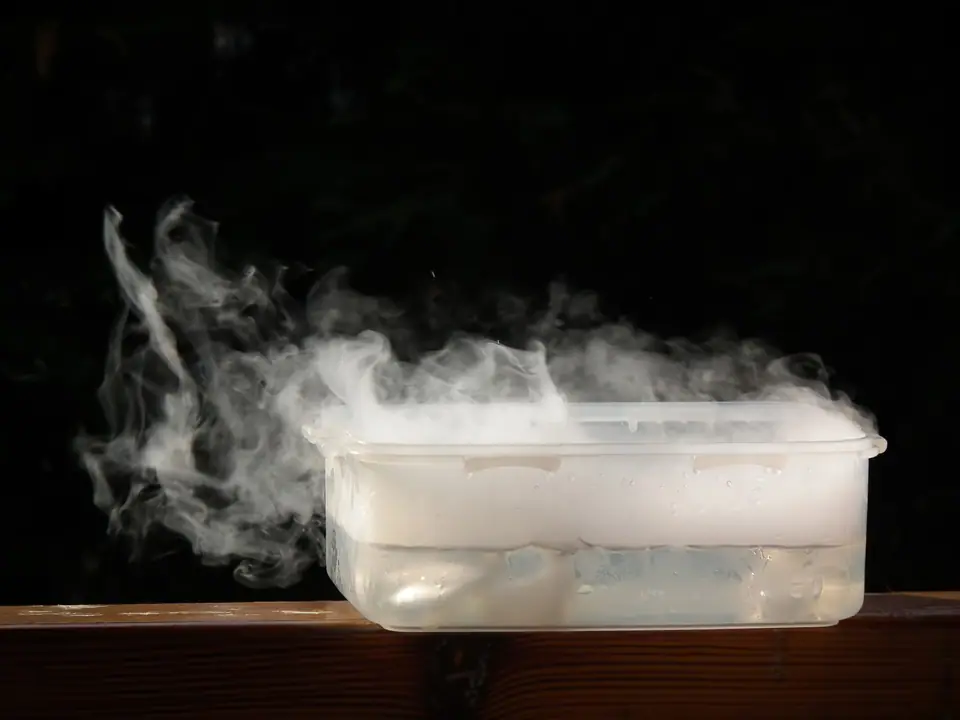 Recipiente de plástico con agua y hielo seco del que se derrama una niebla blanca sobre fondo oscuro