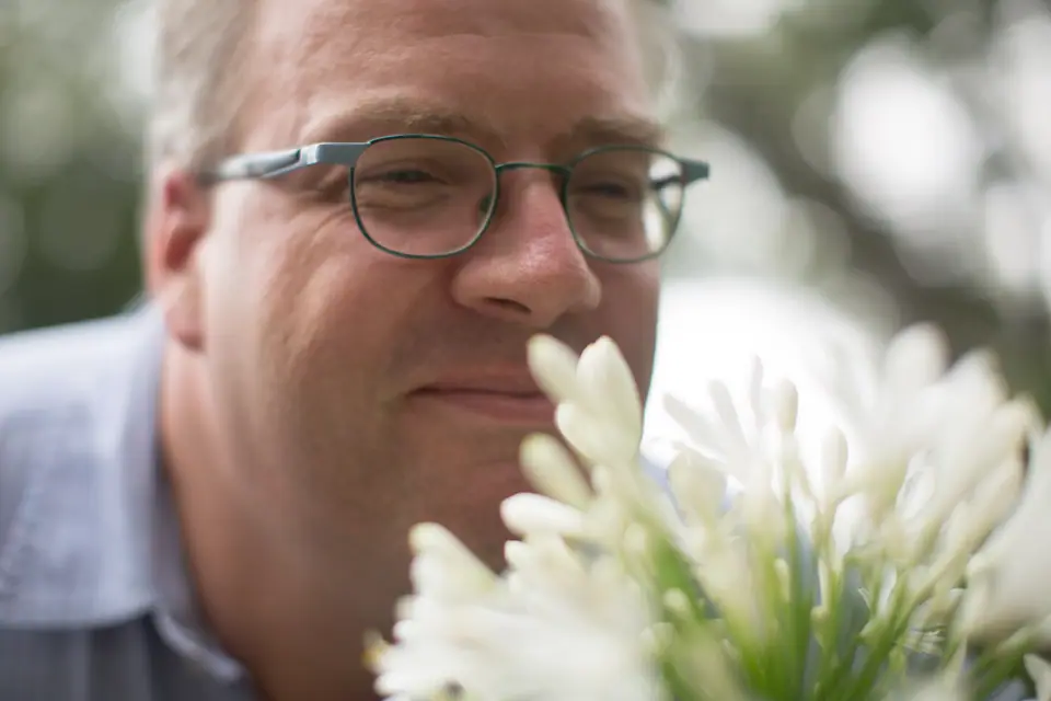 Un hombre de anteojos acerca la nariz a un ramo de flores blancas para olerlas