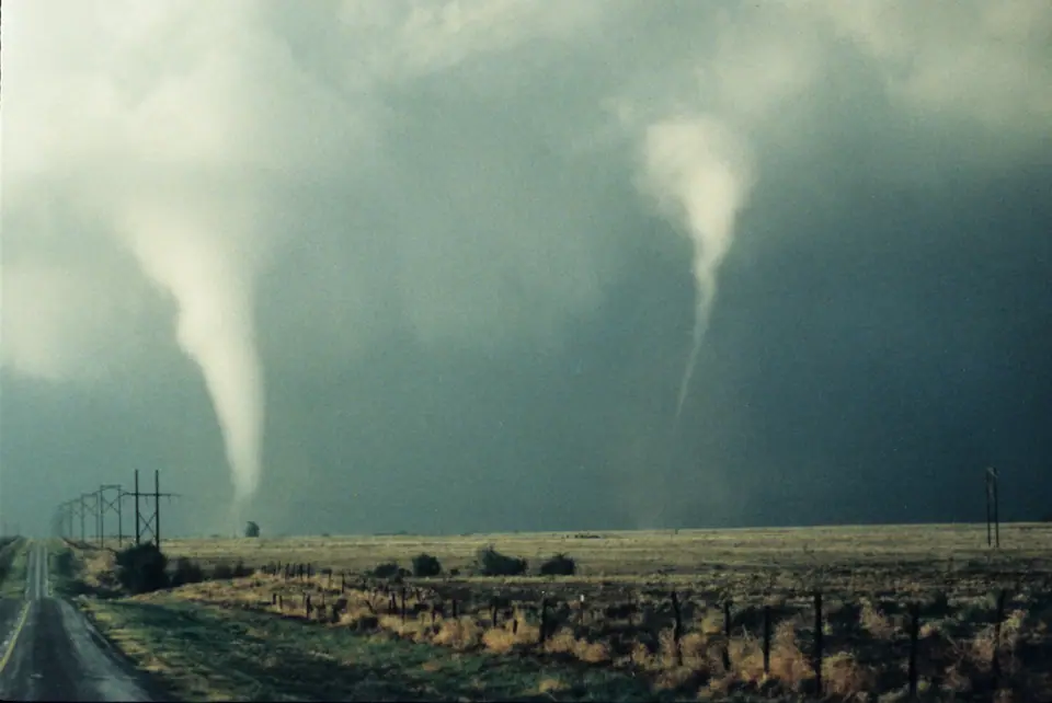 Dos tornados blancos en forma de embudo bajan de una nube oscura sobre una llanura con un camino y postes de luz